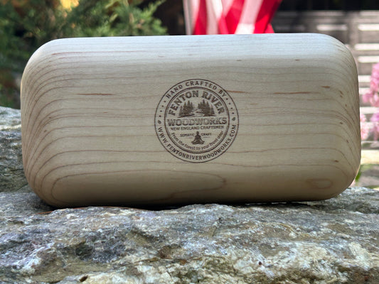 A light natural wood rectangular box with rounded corners and a visible engraved logo reading 'Fenton River Woodworks' resting on a stone surface outdoors.