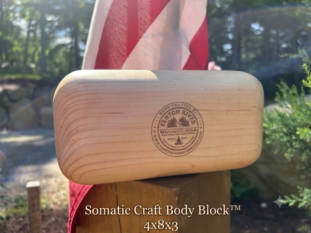 A light natural wood Somatic Craft Body Block with a circular 'Fenton River Woodworks' logo, resting on a wooden post outdoors with a blurred American flag in the background.