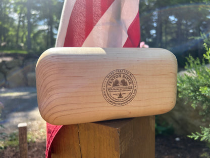 A light natural wood rectangular block with rounded ends, featuring a circular laser-engraved logo for 'Fenton River Woodworks', resting on a wooden post outdoors with an American flag blurred in the background.
