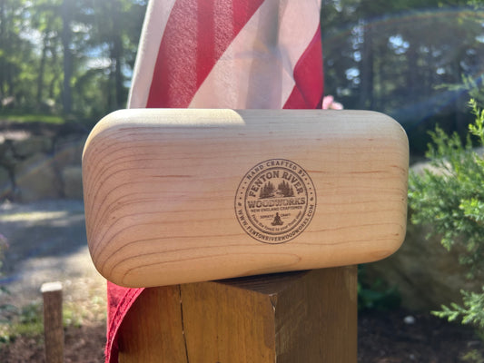 A light natural wood rectangular block with rounded ends, featuring a circular laser-engraved logo for 'Fenton River Woodworks', resting on a wooden post outdoors with an American flag blurred in the background.